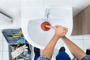 High Angle View Of Male Plumber Using Plunger In Bathroom Sink
